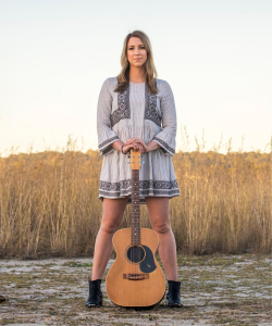 Kaylens Rain standing in a field holding an acoustic guitar
