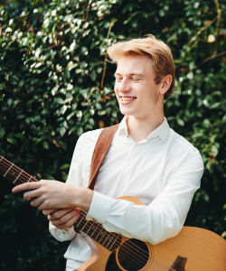 Manning Patston smiling and playing acoustic guitar outdoors