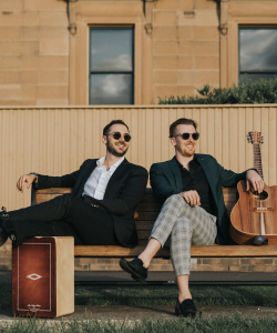 Vale duo in sunglasses seated on a bench with acoustic guitar and cajon