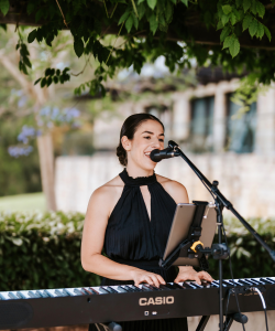 Sarah Vella singing into a microphone and playing keyboard beneath garden foliage.