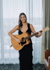 Grace Mae holding an acoustic guitar by a window