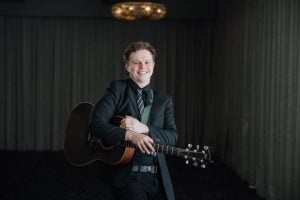 Reuben Ryan smiling with an acoustic guitar, wearing a dark suit