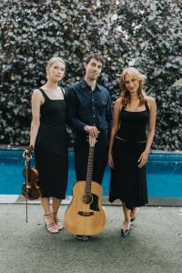 Suade acoustic trio holding violin and guitar in a poolside portrait.